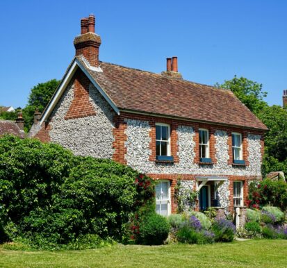 Charming traditional brick cottage with a tiled roof, surrounded by lush greenery and colorful garden flowers under a clear blue sky